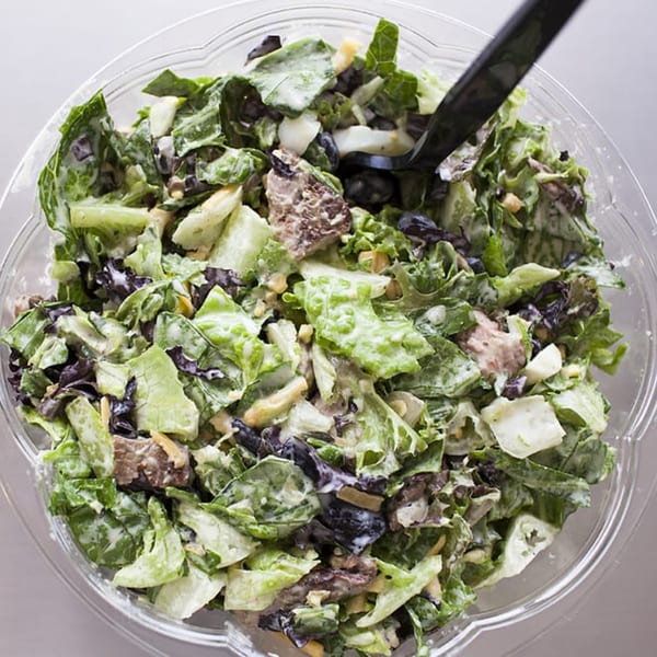 A close-up of a Fourleaf Chopped Salad with mixed greens, black olives, and dressing in a clear bowl.
