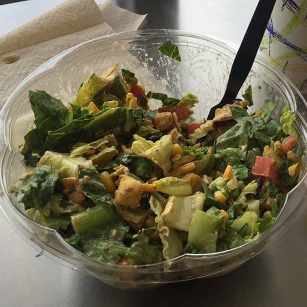 A Fourleaf Chopped Salad in a clear plastic bowl with a black fork, featuring greens, corn, and tomatoes.
